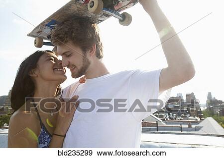 Stock Photo - Mixed Race couple hugging on rooftop with skateboard. Fotosearch
