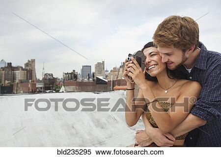Stock Photography - Mixed Race couple hugging on rooftop with video camera. Fotosearch