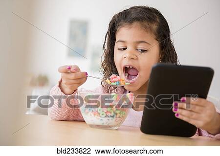 Stock Image - Mixed Race girl eating cereal and using digital tablet. Fotosearch