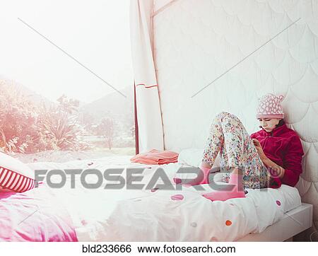 Stock Photograph - Mixed Race girl sitting on bed texting on cell phone. Fotosearch