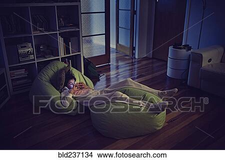 Picture - Mixed Race girl using cell phone laying on bean bags in dark. Fotosearch