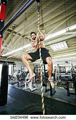 Stock Photograph - Mixed Race man climbing rope in gymnasium. Fotosearch