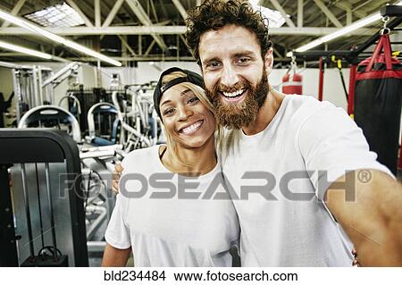 Picture - Mixed Race man couple posing for cell phone selfie in gymnasium. Fotosearch