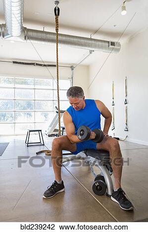 Stock Image - Mixed Race man curling dumbbell in gymnasium. Fotosearch