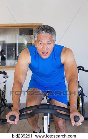 Stock Image - Mixed Race man riding stationary bicycle in gymnasium. Fotosearch