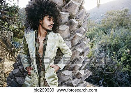 Stock Photo - Mixed Race man wearing jacket leaning on tree trunk. Fotosearch