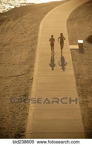Stock Photo - Mixed Race mother and daughter running on path at beach. Fotosearch