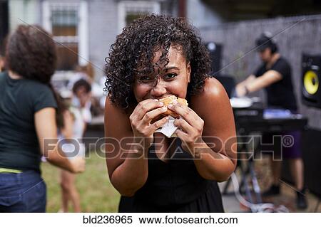 Stock Photography - Mixed Race woman eating hamburger at backyard party. Fotosearch