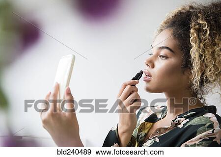 Stock Photo - Mixed Race woman holding cell phone applying lipstick. Fotosearch