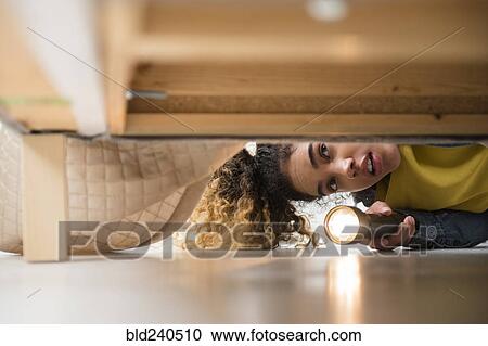 Stock Image - Mixed Race woman looking under bed with flashlight. Fotosearch