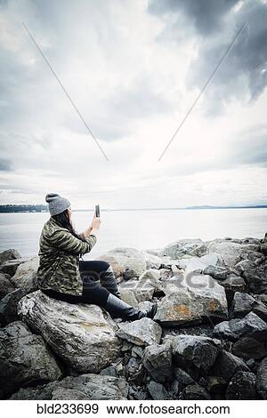 Stock Photo - Mixed Race woman posing for cell phone selfie on rocks at ocean. Fotosearch