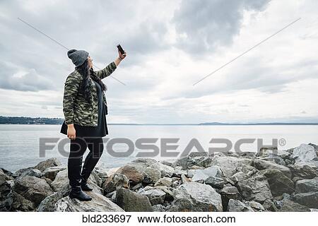 Mixed Race woman standing on rocks at ocean posing for cell phone selfie View Large Photo Image Stock Photo - Mixed Race woman standing on rocks at ocean posing for cell phone selfie. Fotosearch