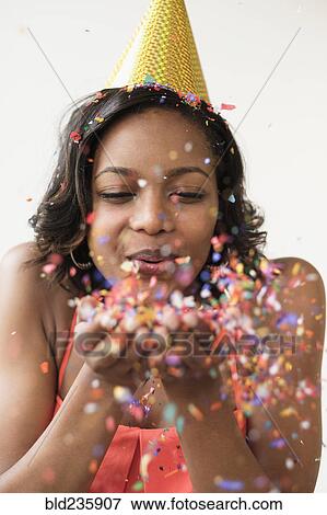 Mixed Race woman wearing party hat blowing confetti View Large Photo Image Stock Photo - Mixed Race woman wearing party hat blowing confetti. Fotosearch