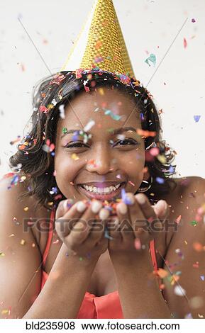 Stock Photo - Mixed Race woman wearing party hat blowing confetti. Fotosearch