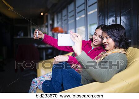 Stock Image - Mother and daughter sitting on sofa posing for cell phone selfie. Fotosearch