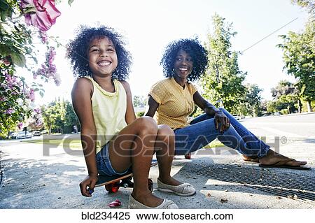 Stock Photography - Mother and daughter sitting on skateboard on sidewalk. Fotosearch