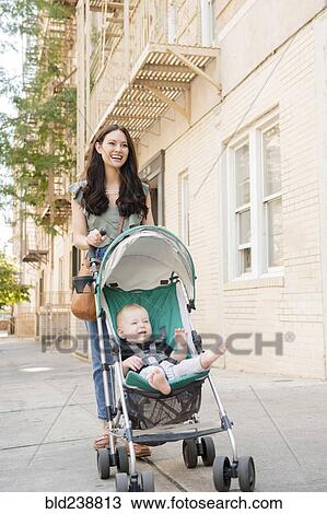 Stock Image - Mother pushing baby son in stroller in city. Fotosearch