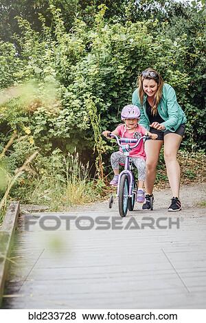 Stock Photo - Mother watching daughter riding bicycle with training wheels. Fotosearch
