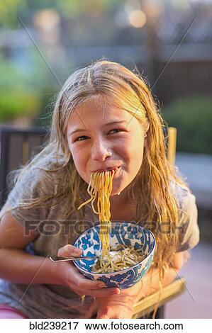 Stock Photo - Noodles hanging from mouth of Caucasian girl holding bowl. Fotosearch