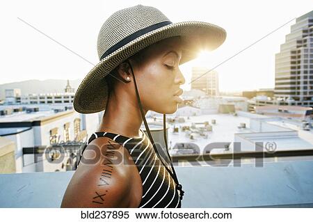 Stock Photography - Pensive Black woman wearing sun hat on rooftop. Fotosearch