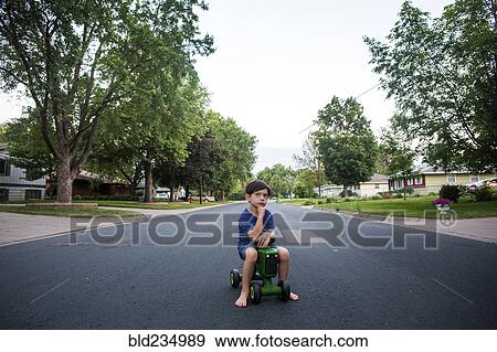 Stock Photo - Pensive mixed race boy sitting on toy tractor in street. Fotosearch