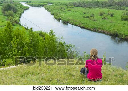 Stock Image - Pensive woman sitting on hill at river. Fotosearch