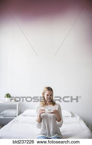 Stock Photograph - Portrait of Caucasian woman sitting on bed drinking tea. Fotosearch