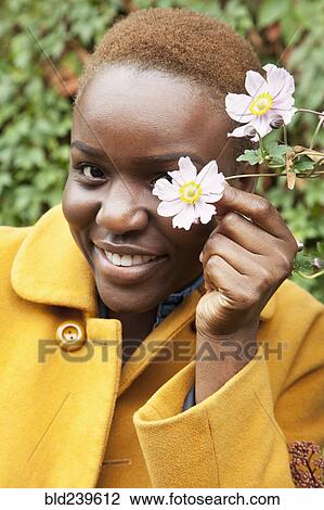 Stock Image - Portrait of smiling Black woman covering eye with flower. Fotosearch