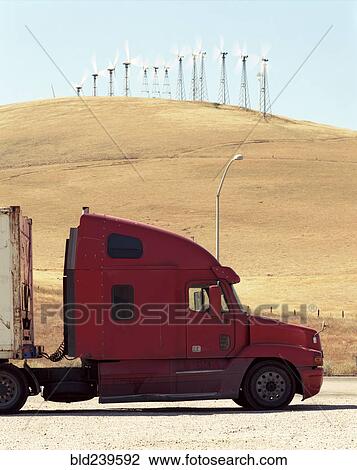 Stock Image - Red semi-truck parked near wind farm. Fotosearch