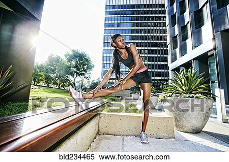 Stock Photography - Serious Black woman stretching leg on urban bench. Fotosearch