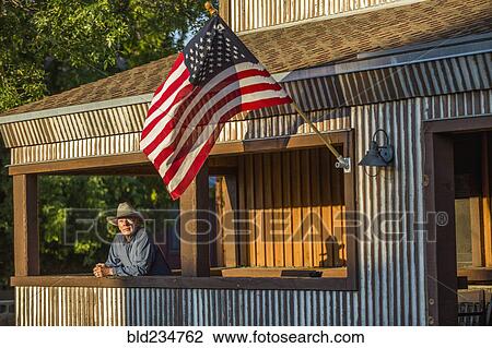 Stock Image - Serious Caucasian farmer on porch with American flag. Fotosearch