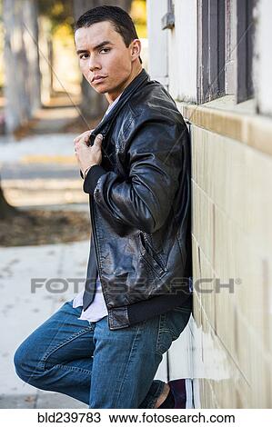 Stock Image - Serious Mixed Race man leaning on wall wearing leather jacket. Fotosearch