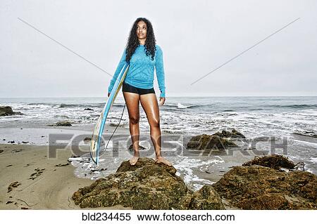 Stock Photography - Serious Mixed Race woman holding surfboards on rocks at beach . Fotosearch