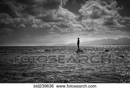 Stock Photograph - Silhouette of Caucasian boy standing on rock in ocean. Fotosearch