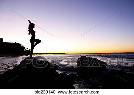 Stock Image - Silhouette of Caucasian woman balancing on rock at beach. Fotosearch