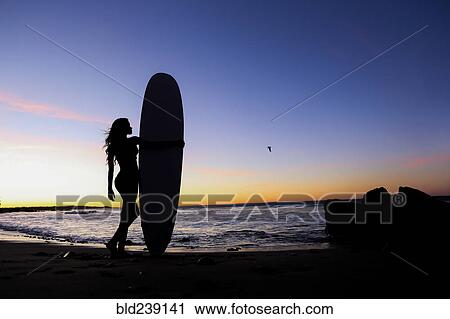 Stock Image - Silhouette of Caucasian woman holding surfboard at beach. Fotosearch