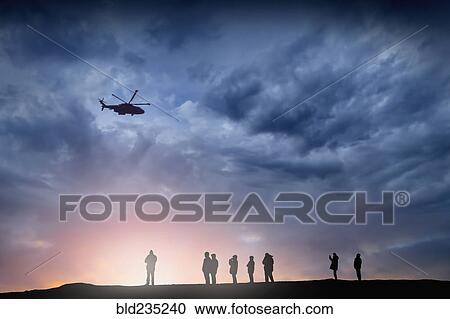 Stock Image - Silhouette of helicopter flying over people at sunset. Fotosearch