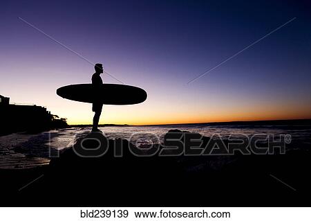 Stock Photo - Silhouette of Hispanic man holding surfboard at beach. Fotosearch
