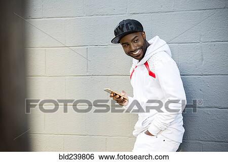 Stock Photograph - Smiling Black man leaning on wall holding cell phone. Fotosearch