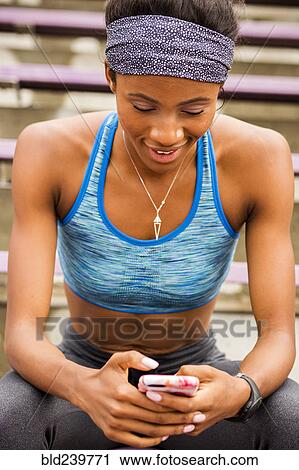 Stock Image - Smiling Black woman sitting on bleachers texting on cell phone. Fotosearch