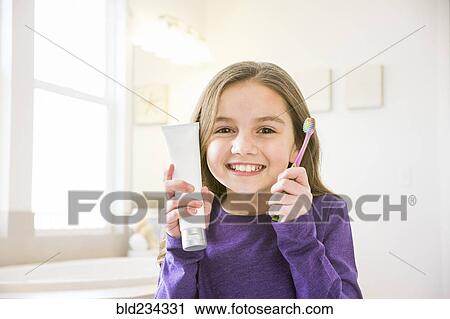 Stock Image - Smiling Caucasian girl holding toothbrush and toothpaste tube in bathroom. Fotosearch
