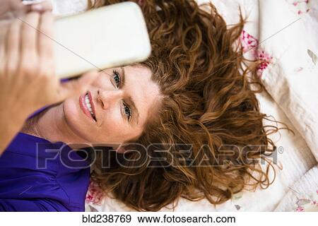 Stock Photo - Smiling Caucasian woman laying on bed texting on cell phone. Fotosearch