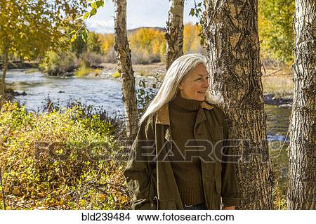 Picture - Smiling Caucasian woman leaning on tree near river. Fotosearch