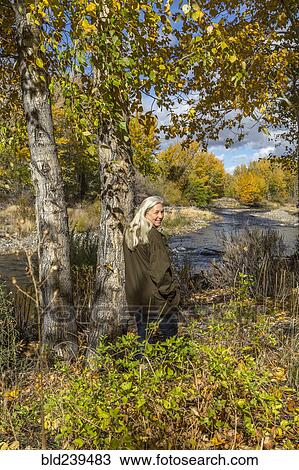 Stock Image - Smiling Caucasian woman leaning on tree near river. Fotosearch