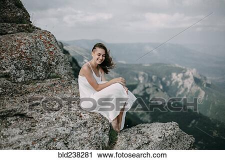 Stock Image - Smiling Caucasian woman sitting on rock overlooking landscape. Fotosearch