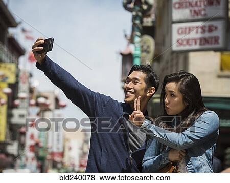 Stock Photo - Smiling Chinese couple posing for cell phone selfie in city. Fotosearch