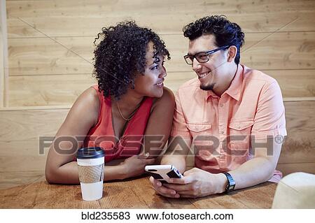 Stock Image - Smiling couple using cell phone in coffee shop. Fotosearch