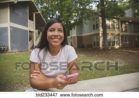 Smiling Hispanic girl holding cell phone near apartments View Large Photo Image Stock Photo - Smiling Hispanic girl holding cell phone near apartments. Fotosearch