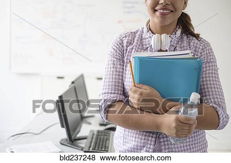 Smiling Hispanic woman holding books and water bottle in computer lab View Large Photo Image Stock Photo - Smiling Hispanic woman holding books and water bottle in computer lab. Fotosearch