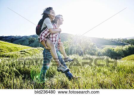 Stock Image - Smiling Man carrying girlfriend piggyback on hill. Fotosearch